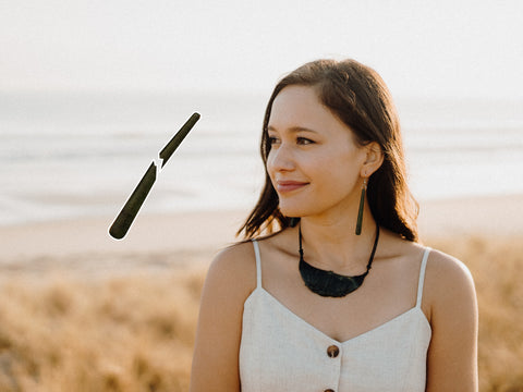 A woman on a beach at dawn wears a greenstone earring and a pounamu necklace. Beside her, a magnified image of a broken earring floats, showing the item's damaged state.