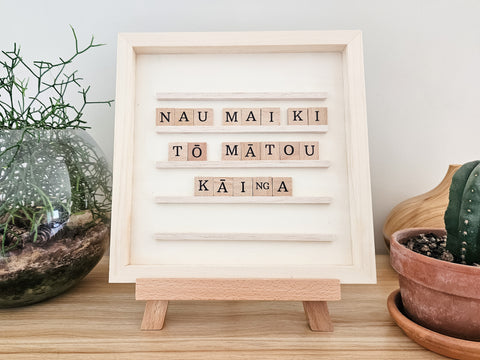 Wooden display board with "Nau mai ki tō mātou kāinga" in wooden letter tiles, alongside indoor plants on a shelf.