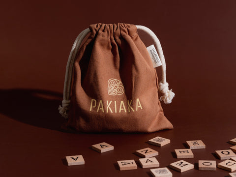 Fabric drawstring bag labelled 'Pakiaka' surrounded by scattered wooden Māori letter tiles on a deep brown background.