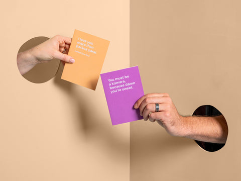 Hands from opposite sides popping through holes exchanging cheeky greeting cards with the texts 'I love you more than Parāoa Parai' and 'You must be a kūmara, because damn you're sweet."