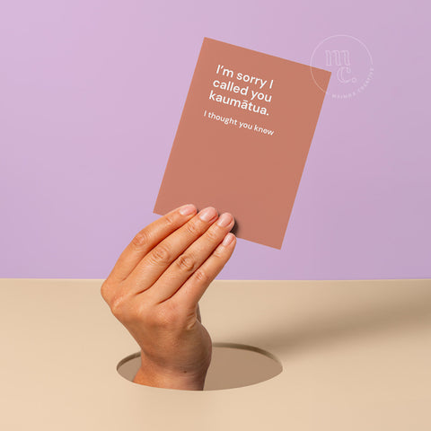 Hand peeking out of hole holding a birthday greeting card with the text 'I’m sorry I called you kaumātua. I thought you knew,' against a pastel purple and beige background.