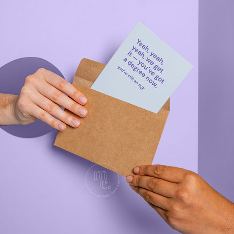 Two hands passing and receiving a humorous graduation greeting card poking out of a kraft envelope, with the message 'Yeah yeah yeah we get it—you’ve got a degree now,' on a pastel purple background