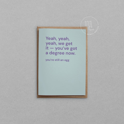 Greeting card with text 'Yeah, yeah, yeah, we get it — you’ve got a degree now. You’re still an egg' on a bright pastel blue background, placed on a light surface