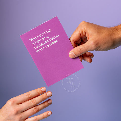 Hand passing a cute greeting card with the text 'You must be a kūmara, because damn you’re sweet' on a pastel purple background