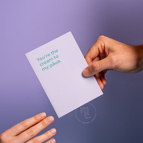 Hand passing a cute greeting card with the text 'You’re the cream to my pāua.' on a pastel purple background
