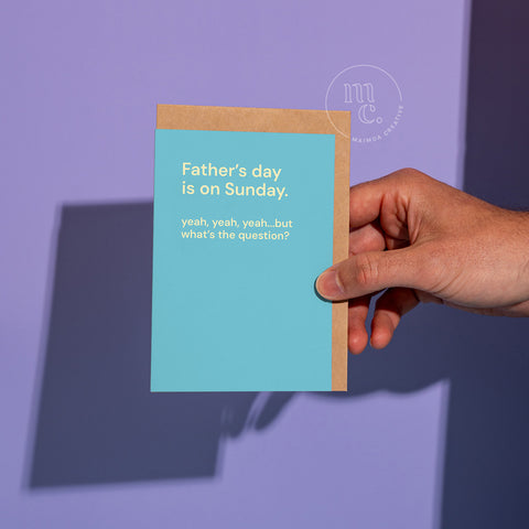 Hand holding a funny Father’s Day card on a purple background with the quote “Father’s day is on Sunday. yeah, yeah, yeah…but what’s the question?” by Maimoa Creative.