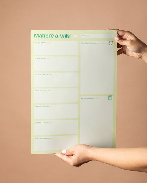 Person holding a green magnetic weekly planner with 'Mahere ā-wiki' text on a brown background
