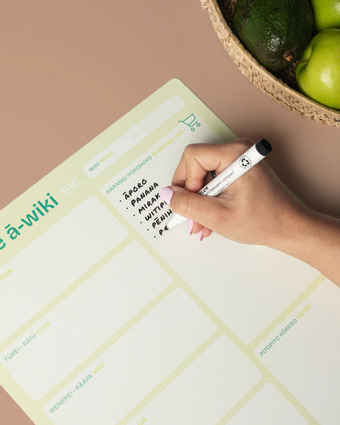 Hand writing a grocery list on a green magnetic weekly planner with a marker, next to a bowl of avocados and green apples