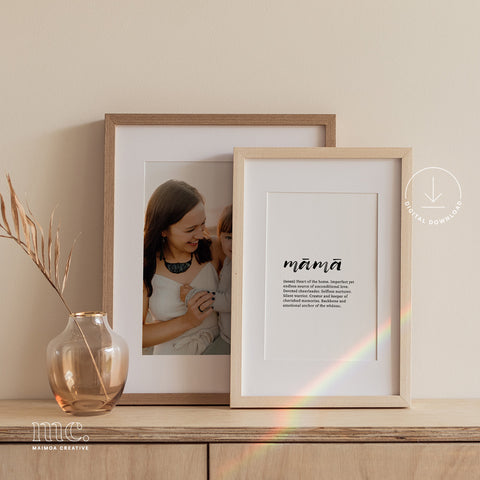Two framed prints on a sideboard; one with a 'māmā' definition, one a mother-child photo, with a vase and dried stems.