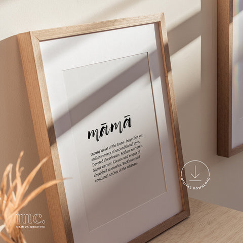 Framed 'māmā' definition print on a sideboard beside a vase with dried branches, casting shadows.