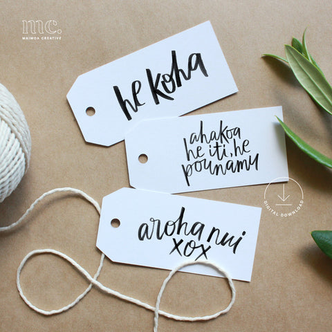 Three white gift tags with black Māori text on a brown surface with string and leaves.