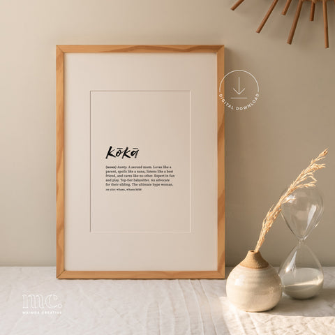 Hand-lettered 'kōkā' definition print in a light wood frame, next to a vase and hourglass on a beige surface.