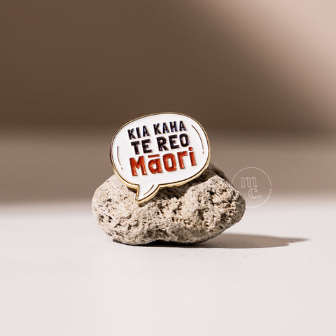  An enamel pin in a speech bubble shape with the inscription "Kia Kaha Te Reo Māori" placed on a textured pumice stone, with clear white, red, and black colours and brass detailing around the edges.