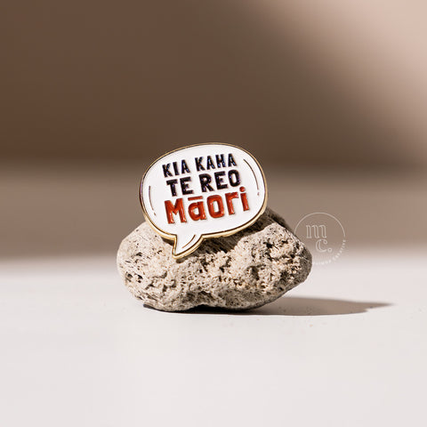 An enamel pin in a speech bubble shape with the inscription "Kia Kaha Te Reo Māori" placed on a textured pumice stone, with clear white, red, and black colours and brass detailing around the edges.