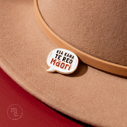 An enamel badge pin with the words "Kia Kaha Te Reo Māori" written in bold letters on a tan wool hat