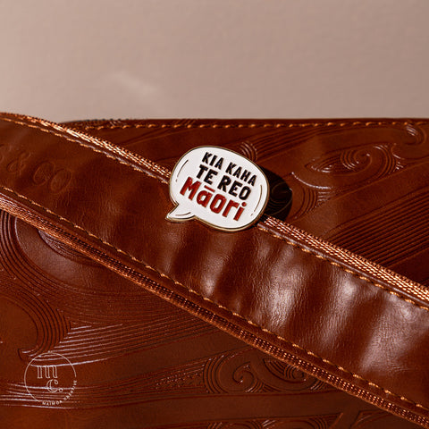 An enamel badge pin with the words "Kia Kaha Te Reo Māori" written in bold letters on a brown leather bag with Māori motifs and designs