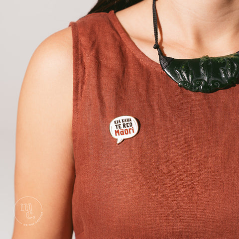 An enamel badge pin with the words "Kia Kaha Te Reo Māori" written in bold letters on a rust colour dress worn by a Māori woman wearing a taonga