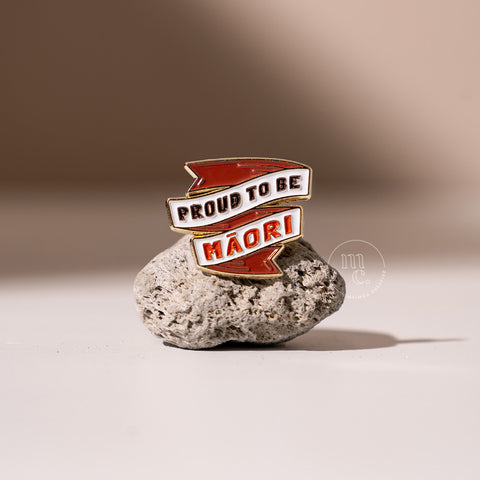 An enamel pin in a banner shape with the inscription "Proud to be Māori" placed on a textured pumice stone, with clear white, red, and black colours and brass detailing around the edges.