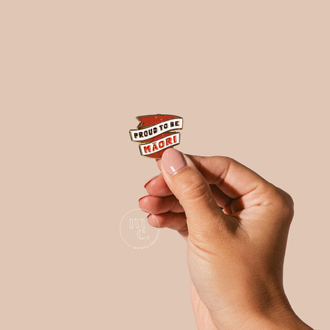 A hand holding an enamel pin with the words "Proud to be Māori" written in bold letters against a neutral-toned background.