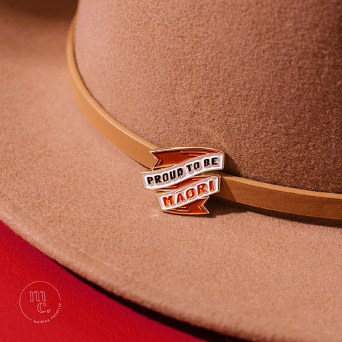 An enamel badge pin with the words "Proud to be Māori" written in bold letters on a tan wool hat