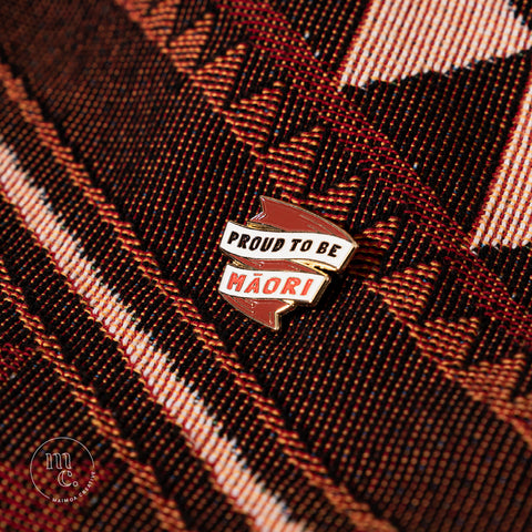 An enamel pin with the inscription "Proud To Be Māori" placed on a cotton red and orange blanket with Māori designs and patterns, with clear white, red, and black colours and brass detailing around the edges.