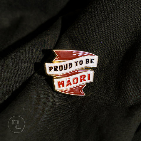 An enamel badge pin with the words "Proud to be Māori" written in bold letters on dark forest green material