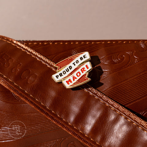An enamel badge pin with the words "Proud to be Māori" written in bold letters on a brown leather bag with Māori motifs and designs