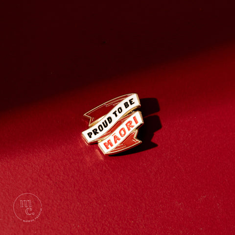 An enamel pin with the inscription "Proud To Be Māori" placed on a bold red background, with clear white, red, and black colours and brass detailing around the edges.