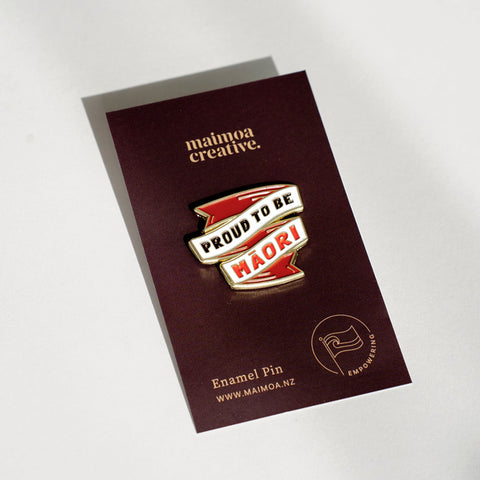 An enamel badge pin with the words "Proud to be Māori" written in bold letters on a dark brown backing card