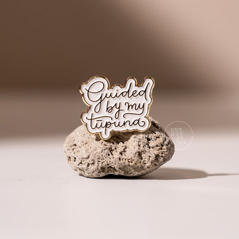 An enamel pin with the inscription "Guided by my Tūpuna" placed on a textured pumice stone, with clear white and brass colours and brass detailing around the edges.