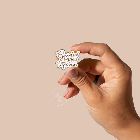 A hand holding an enamel pin with the words "Guided By My Tūpuna" written in gold letters against a neutral-toned background.