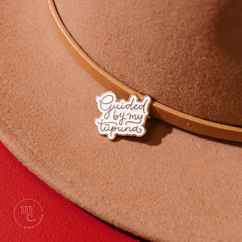 An enamel badge pin with the words "Guided by my Tūpuna" written in gold letters on a tan wool hat