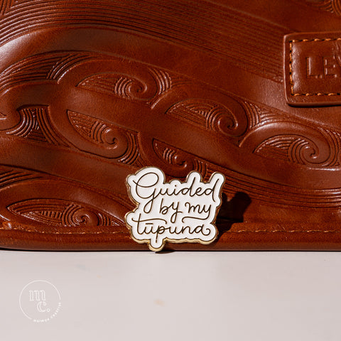 An enamel badge pin with the words "Guided by my Tūpuna" written in gold letters on a brown leather bag with Māori motifs and designs
