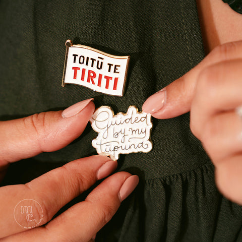 Two hands securing an enamel pin with the words "Guided By My Tūpuna" written in gold letters onto a dark forest green dress with another pin next to it saying "Toitū Te Tiriti"