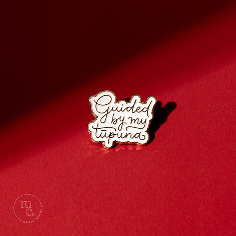 An enamel pin with the inscription "Guided By My Tūpuna" placed on a bold red background, with clear white and gold colours and brass detailing around the edges.