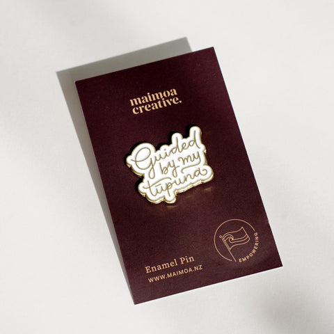 An enamel badge pin with the words "Guided by my Tūpuna" written in gold letters on a dark brown backing card
