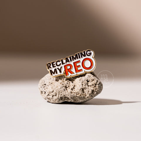 An enamel pin with the inscription "Reclaiming my Reo" placed on a textured pumice stone, with clear white, red, and black colours and brass detailing around the edges.