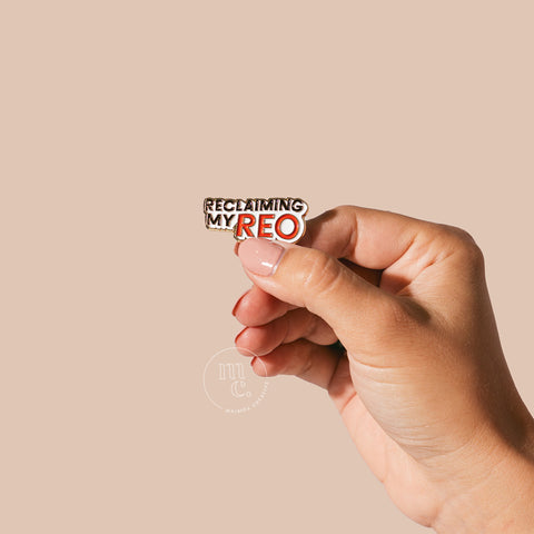 A hand holding an enamel pin with the words "Reclaiming My Reo" written in bold letters against a neutral-toned background.