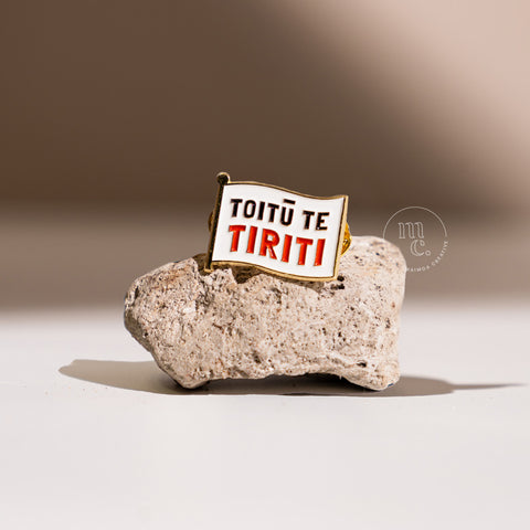 An enamel pin with the inscription "Toitū Te Tiriti" placed on a textured pumice stone, with clear white, red, and black colours and brass detailing around the edges.