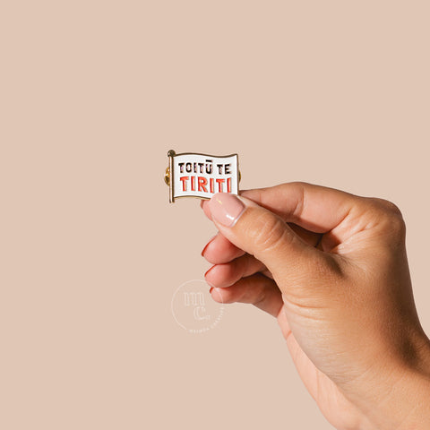 A hand holding an enamel pin with the words "Toitū Te Tiriti" written in bold letters against a neutral-toned background.