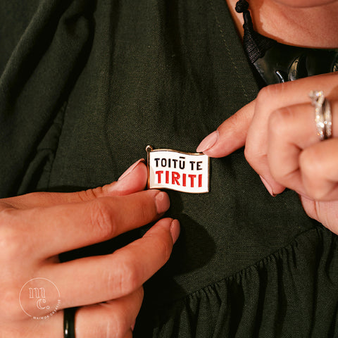 Two hands securing an enamel pin in the shape of a flag with the words "Toitū te Tiriti" written in bold letters onto a dark forest green dress
