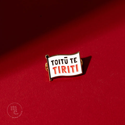 An enamel pin with the inscription "Toitū Te Tiriti" placed on a bold red background, with clear white, red, and black colours and brass detailing around the edges.