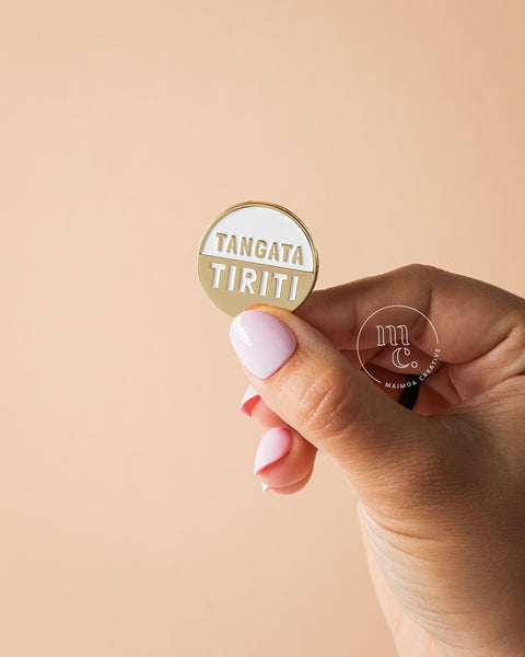 Hand holding a gold and white 'Tangata Tiriti' pin against a beige background