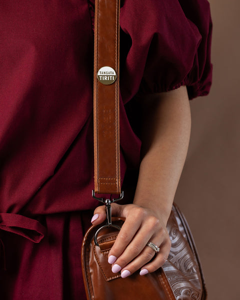 Person holding a brown leather bag with a 'Tangata Tiriti' gold pin wearing a dark plum outfit