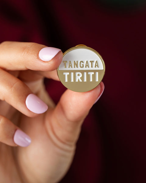 Hand holding a gold and white badge with 'TANGATA TIRITI' text against a blurred background