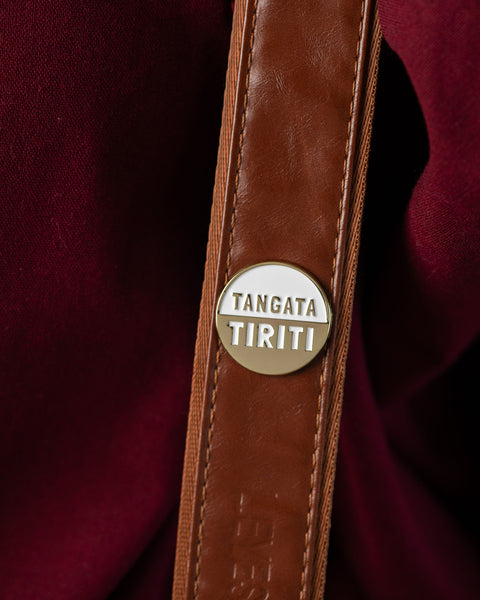 Brown leather strap with a gold and white 'Tangata Tiriti' badge pin on a plum background