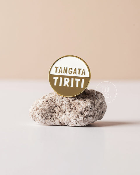 A gold 'Tangata Tiriti' badge pin on a pumice rock with a beige background