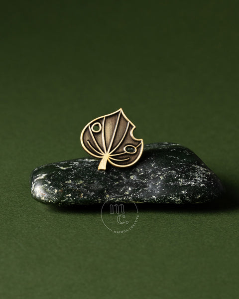 Gold kawakawa leaf pin on a dark stone pounamu with a green background