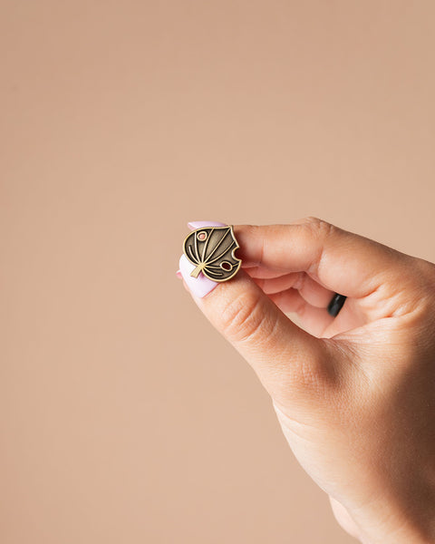 Hand holding a small kawakawa leaf pin against a beige background
