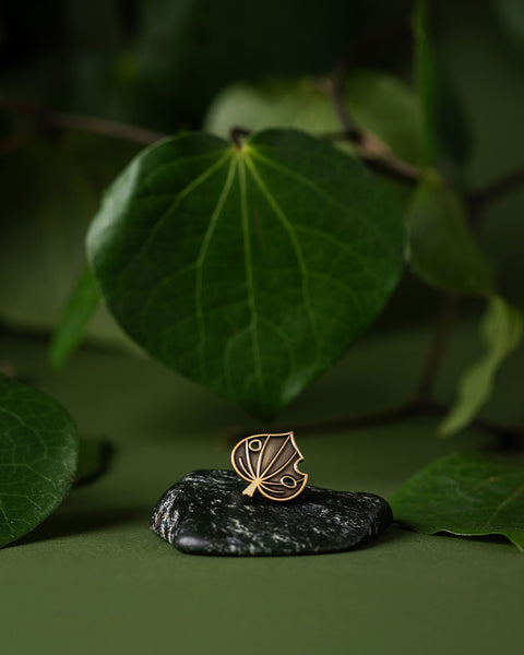 Gold kawakawa leaf pin on a dark stone with green kawakawa leaves in the background